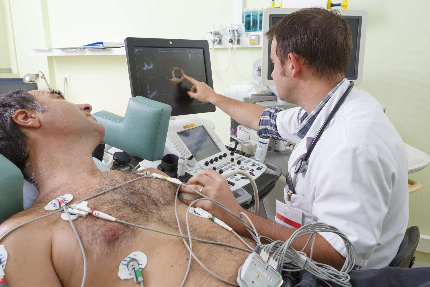 A doctor analyzes echocardiogram results on a monitor while a patient lies in a medical setting connected to monitoring equipment.