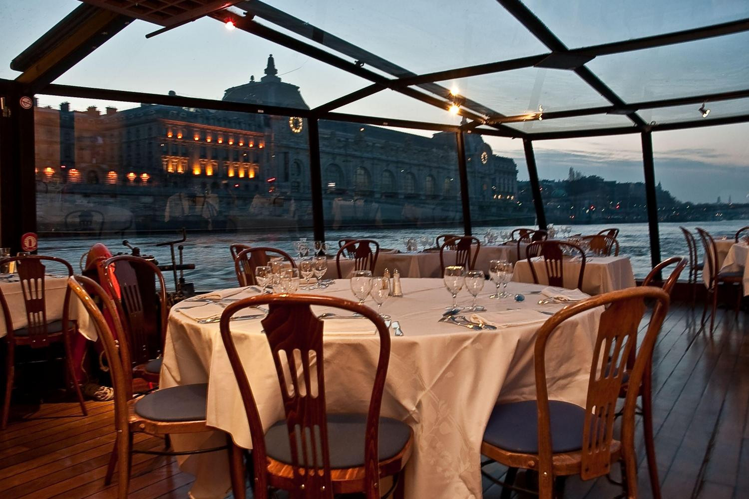 Elegant indoor dining setup on a glass-covered boat, with views of illuminated city buildings and a serene river at dusk.