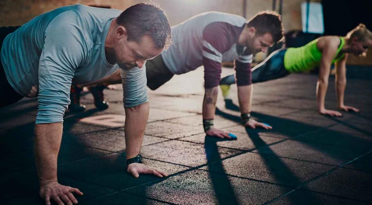 Three individuals perform push-ups on a black rubber gym floor, showcasing a determined workout atmosphere in soft lighting.