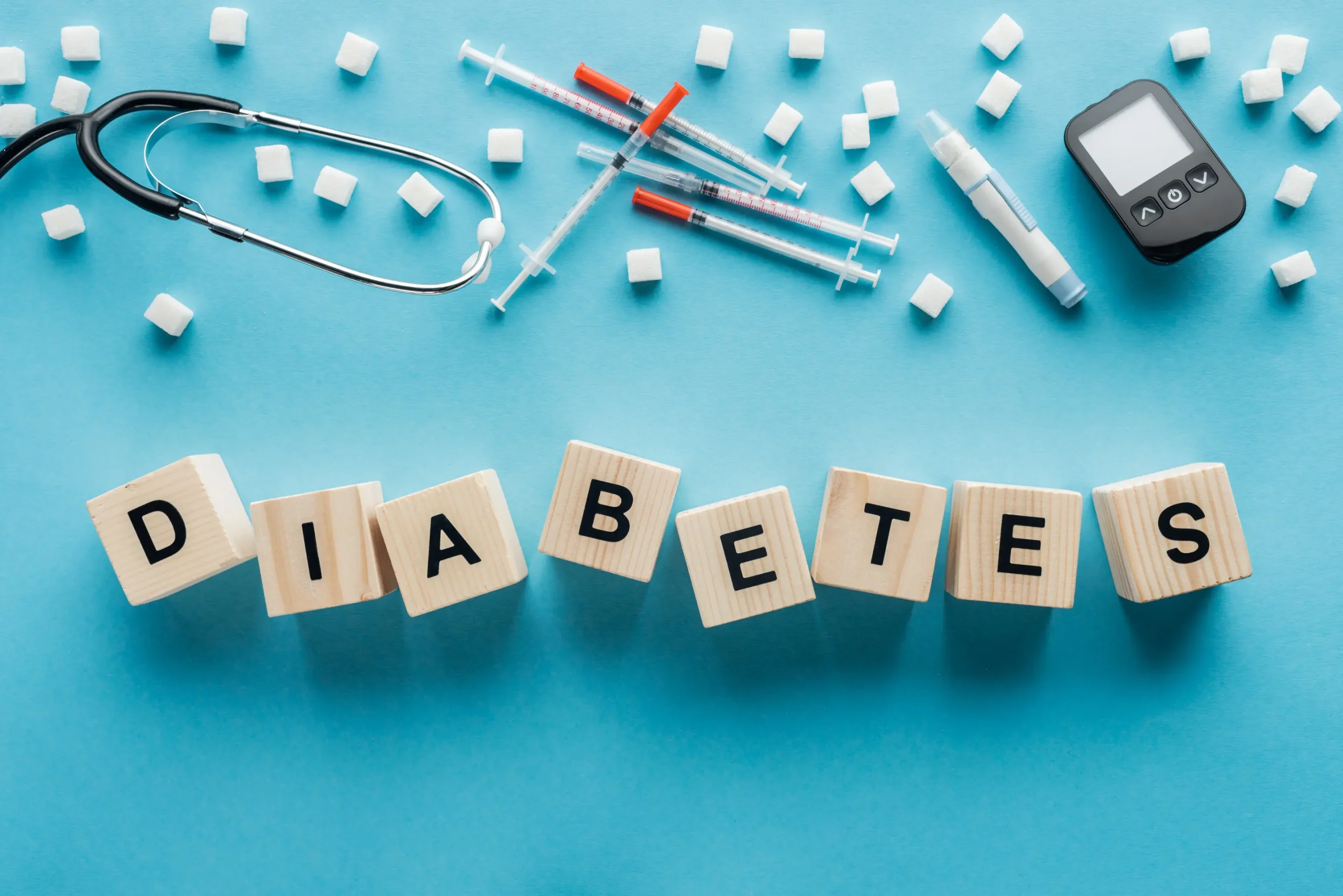 Blocks spelling "DIABETES" surrounded by syringes, a glucose meter, a stethoscope, and sugar cubes on a blue background.