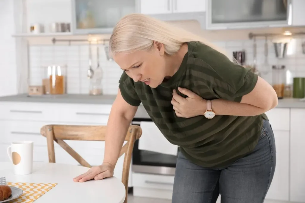 A Woman in a green striped shirt leans on a table, appearing to be in discomfort in a modern kitchen setting. Cardiologist Clinic in Dubai