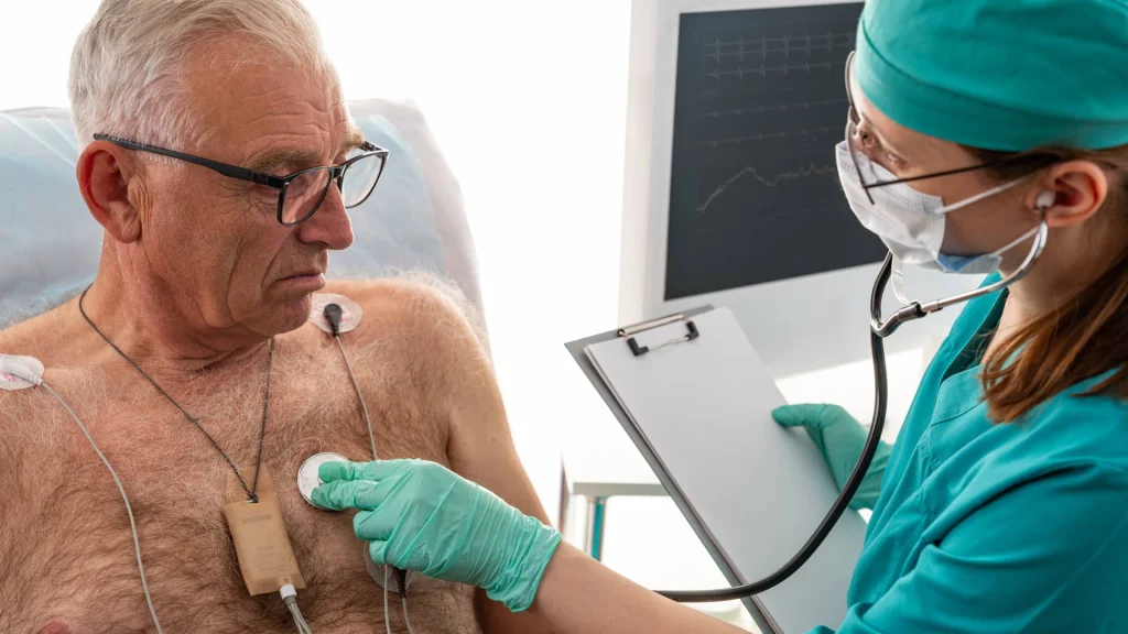 A healthcare professional in teal scrubs and a mask uses a stethoscope on an elderly patient’s chest, with medical equipment in the background.