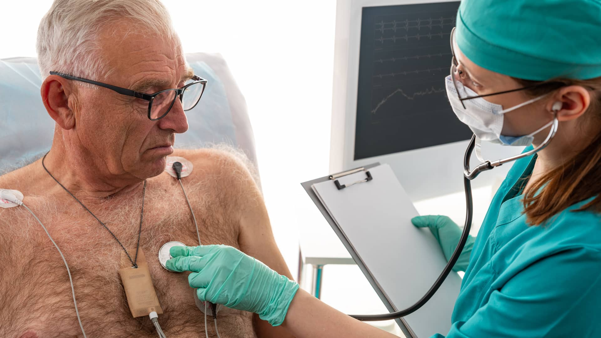 A healthcare professional in teal scrubs and a mask uses a stethoscope on an elderly patient’s chest, with medical equipment in the background.