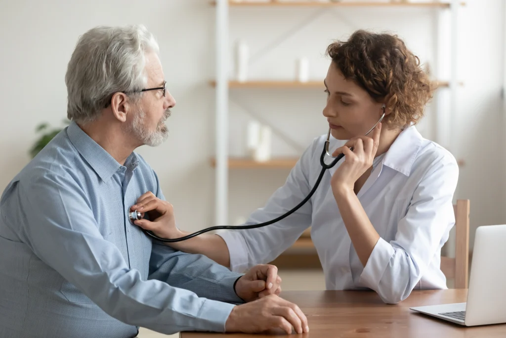 A healthcare professional listens to a patient's heart using a stethoscope in a well-lit office setting. A laptop is on the table.