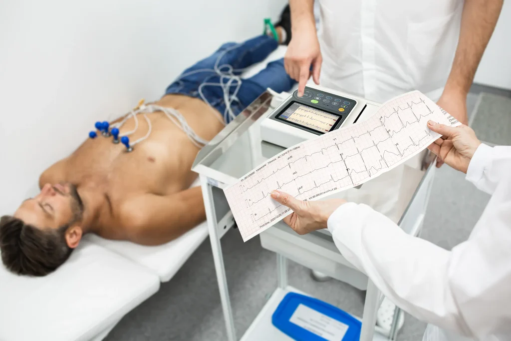 A medical professional holds an ECG printout, while another operates an ECG machine beside a patient connected with leads. check stress