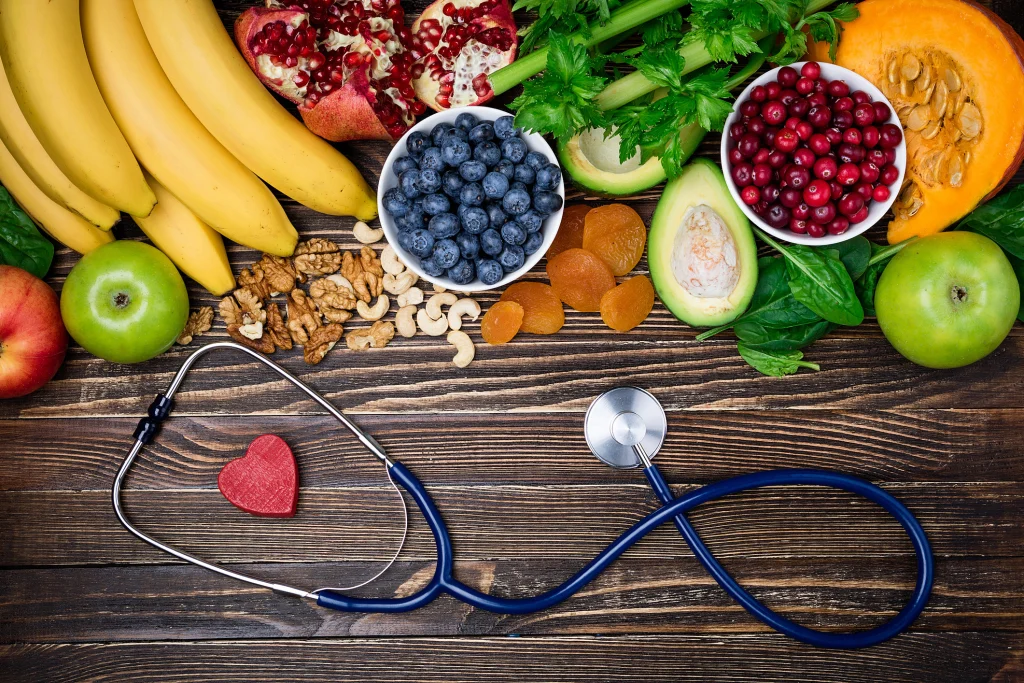 Best cardiologist recommend a variety of fresh fruits, nuts, and vegetables arranged alongside a stethoscope and a red heart shape, symbolizing health and nutrition.