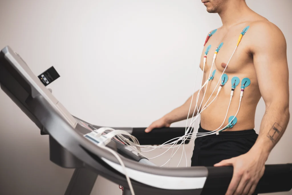 A man undergoing a stress test runs on a treadmill with chest electrodes connected to heart monitoring equipment.
