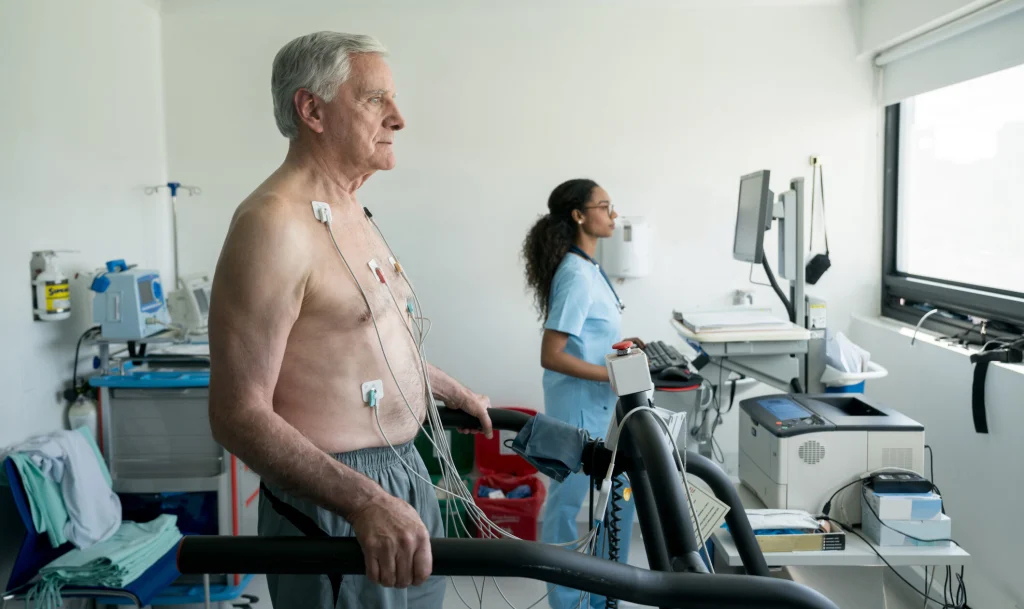 An elderly man undergoes a stress test on a treadmill while a nurse monitors his heart data on a computer in a medical examination room.