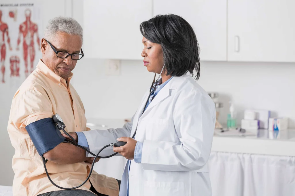 A doctor in a white coat checks an older man's High blood pressure in a medical office, conveying care and professionalism. Medical equipment is visible.