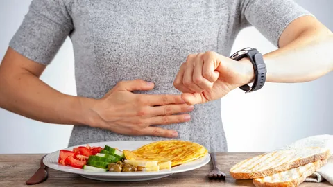 A person in a gray shirt checks their watch while resting a hand on their stomach, next to a plate of food — representing intermittent fasting and heart health in Dubai.