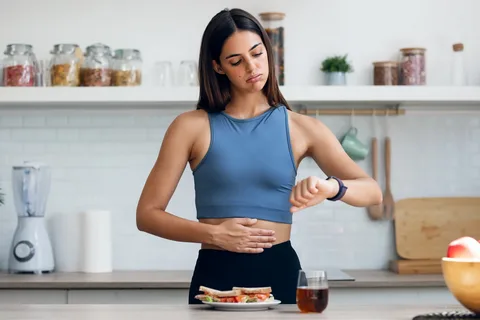 A woman in a kitchen wearing a blue top looks at her watch with a concerned expression, hand on her stomach, with a sandwich and juice in front of her — illustrating intermittent fasting for heart health in Dubai.